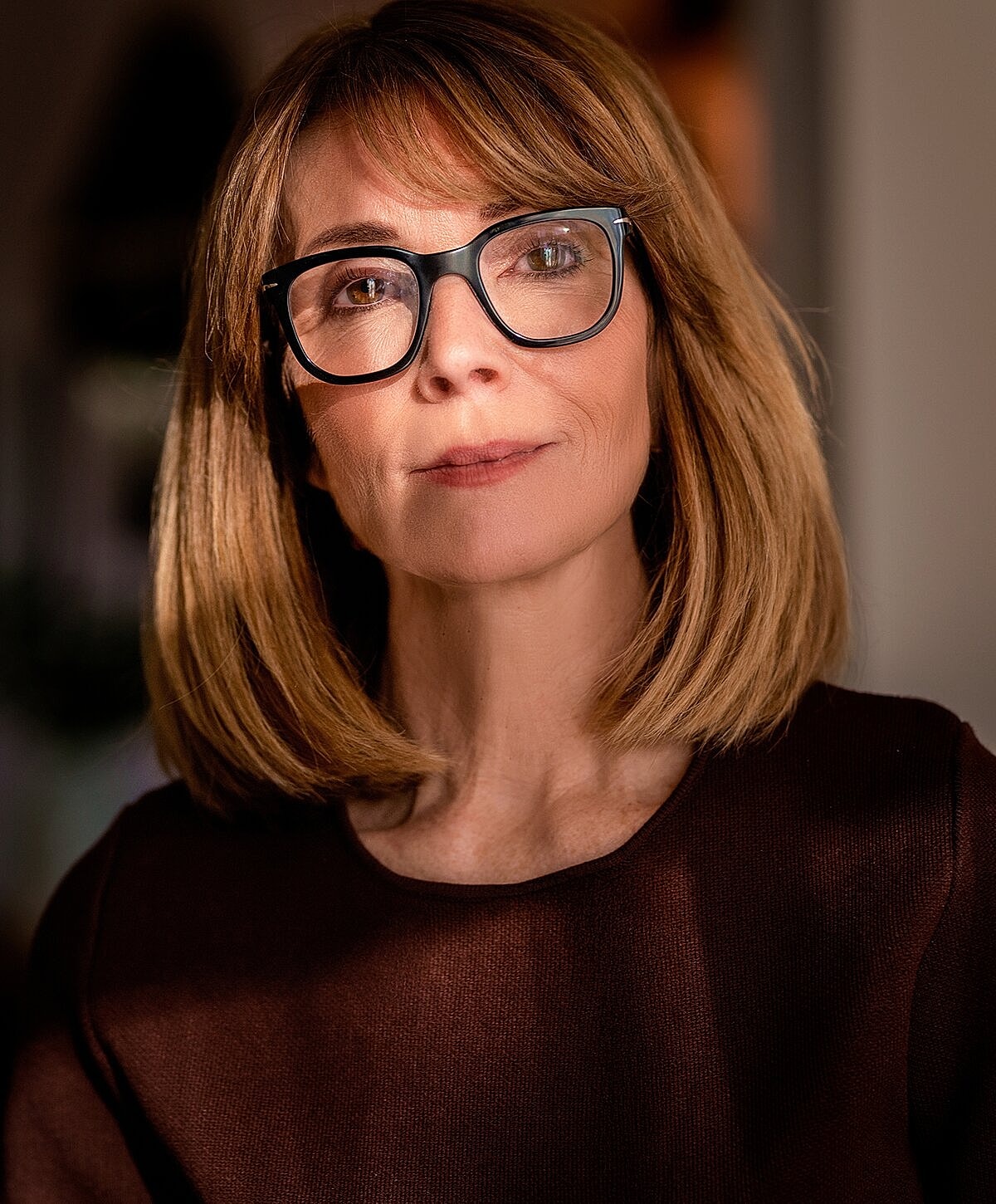 Woman with glasses, warmly lit, thoughtful expression.
